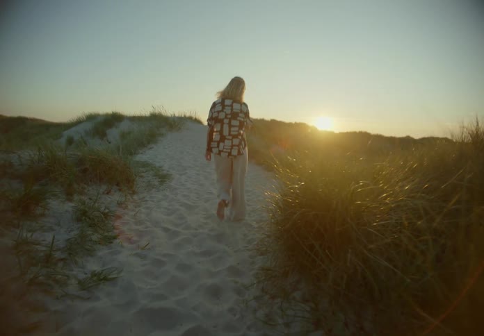 Blonde woman walks along sloping sandy path toward sun sitting on the horizon