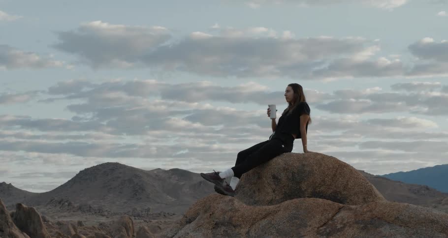 A woman drinks from a thermal mug while sitting atop a rock formation in the badlands during the day