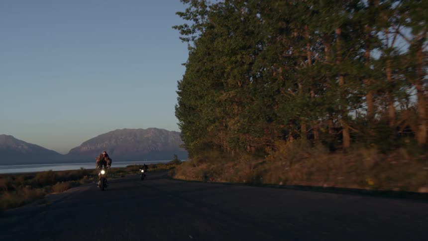 Motorcycles are riding near the water at sunset as one man stands