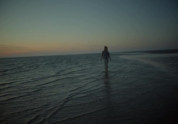 A woman is walking in the water at the edge of the shore at dusk