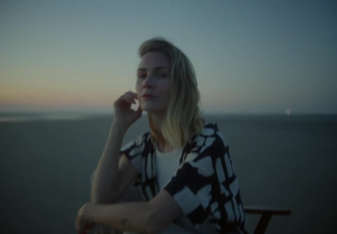 A woman sitting in a chair on the beach turns to look into the camera as it approaches