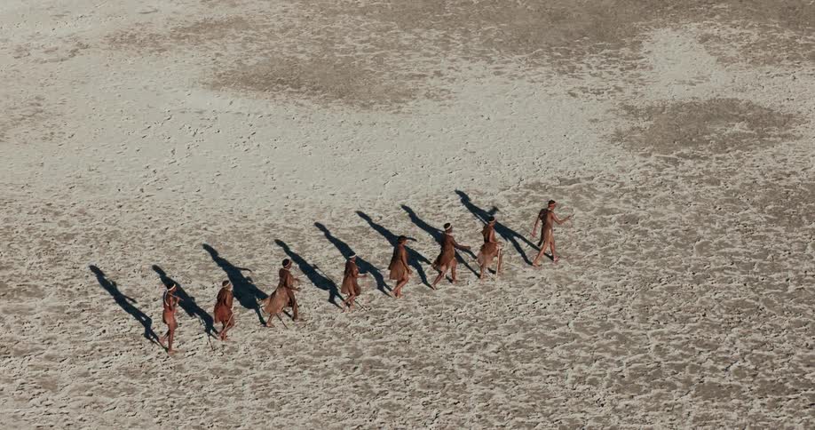 Aerial footage shows a group of bushmen walk in a line along a dusty plain