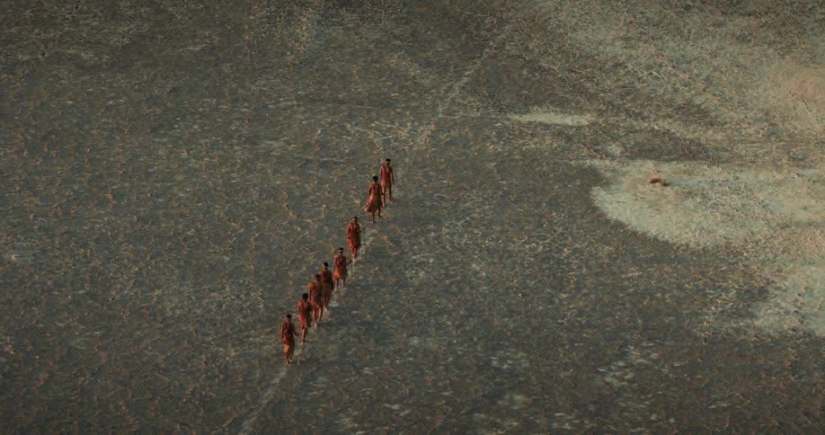 A small group of bushmen walk in a single file line across a salt pan