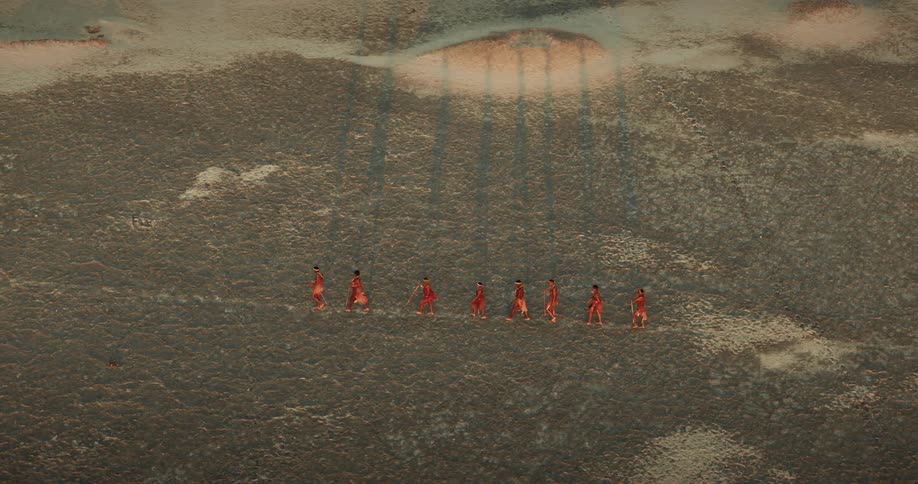 Aerial footage shows a group of bushmen walk in a straight line across barren plain