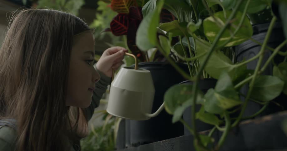A young girl uses a small watering can to water several potted plants