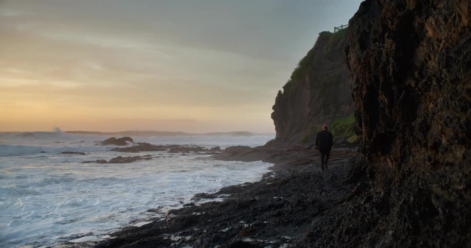 A person walks along a narrow beach beneath a cliff on the coast on a cloudy evening