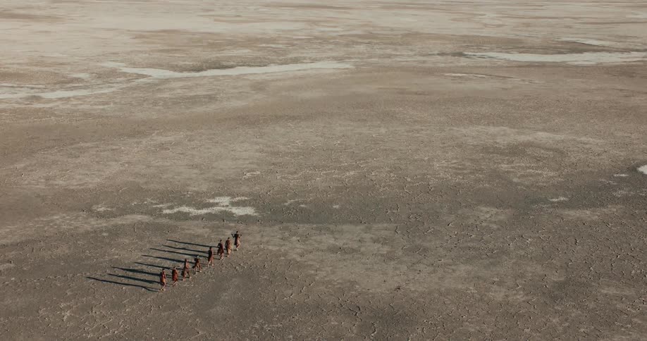 A small group of bushmen walk in a single file line across a wide open plain
