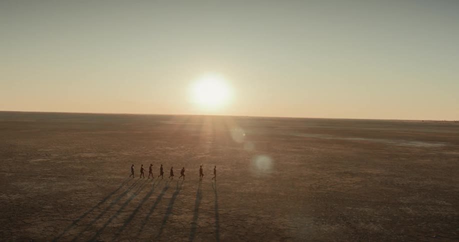 A small group of bushmen walk in a single file line through an arid landscape