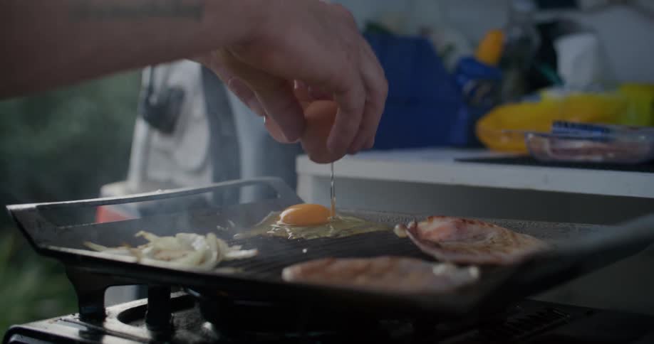 A man cracks an egg over a hot grill outdoors during the day