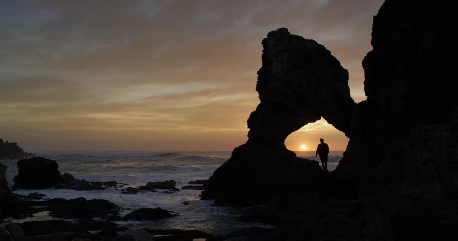 A person stands on a rocky outcrop while watching the sun set over the ocean