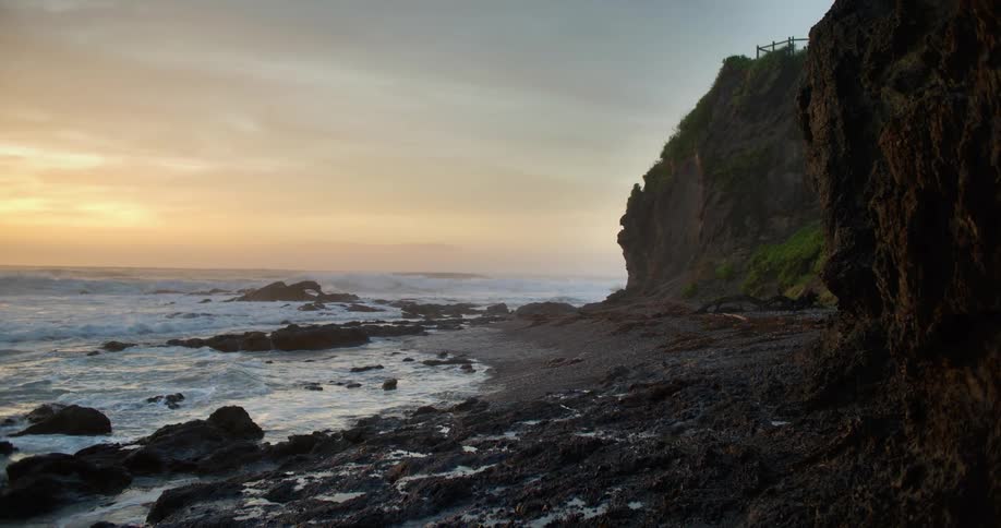 Waves splash on the rocks and roll into shore near a promontory as the sun sets in a cloudy sky