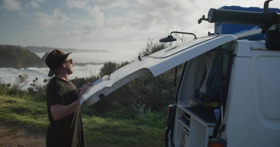 A man wearing a hat is opening the trunk of his van that is parked near a lake experiencing minimal wind