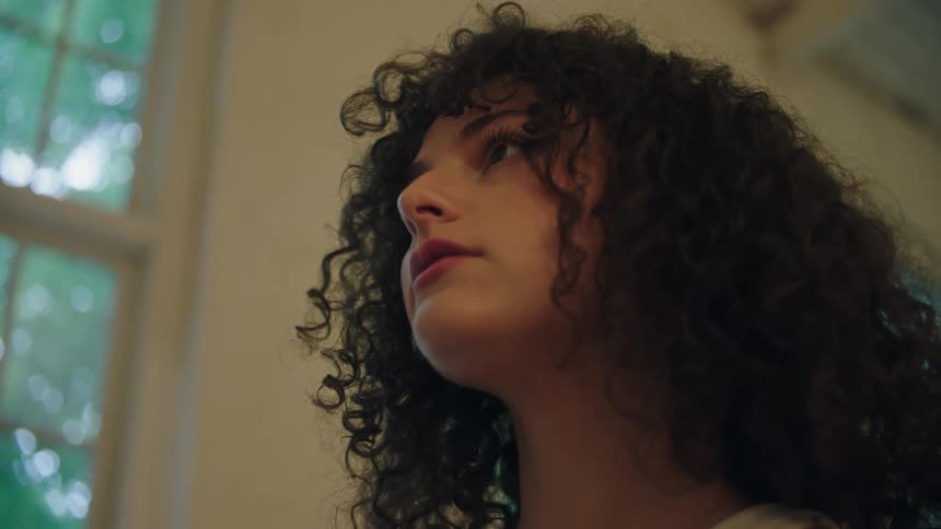 A young woman with curly hair holds a framed picture while looking up at a wall