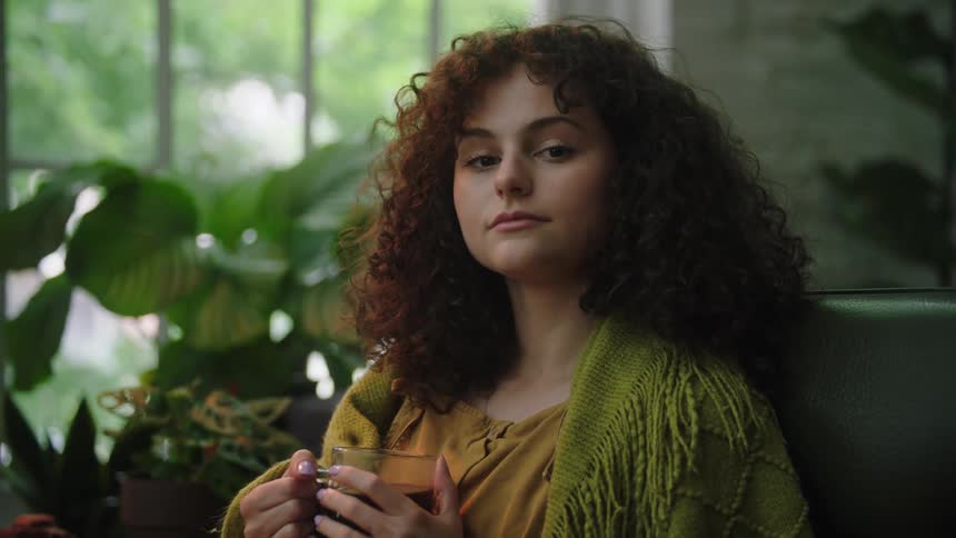 A young woman with curly hair poses while holding a glass mug in her hands