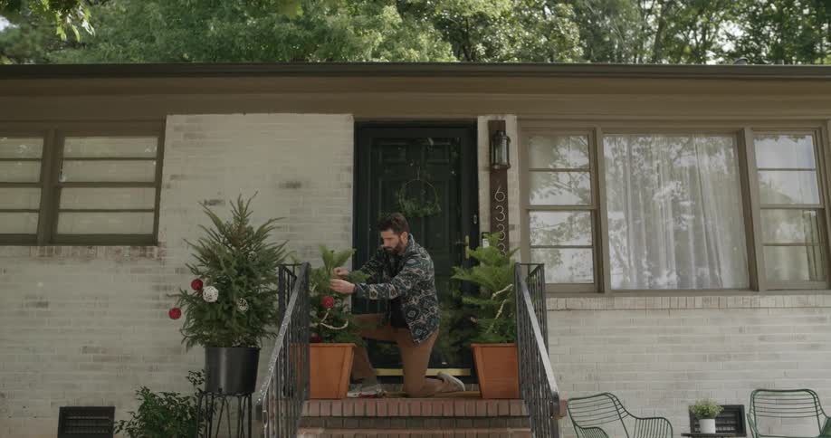 Man kneeling hanging christmas decoration on potted plants in front of a house