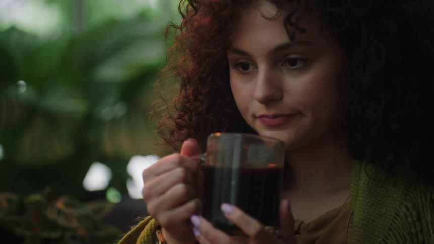 A young woman slowly takes a sip from her coffee mug while looking contemplative