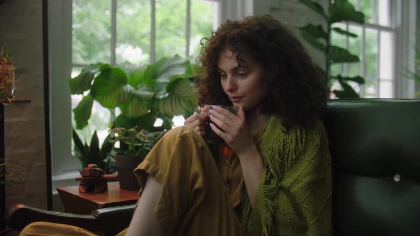 A young woman sits on a couch while holding a hot beverage