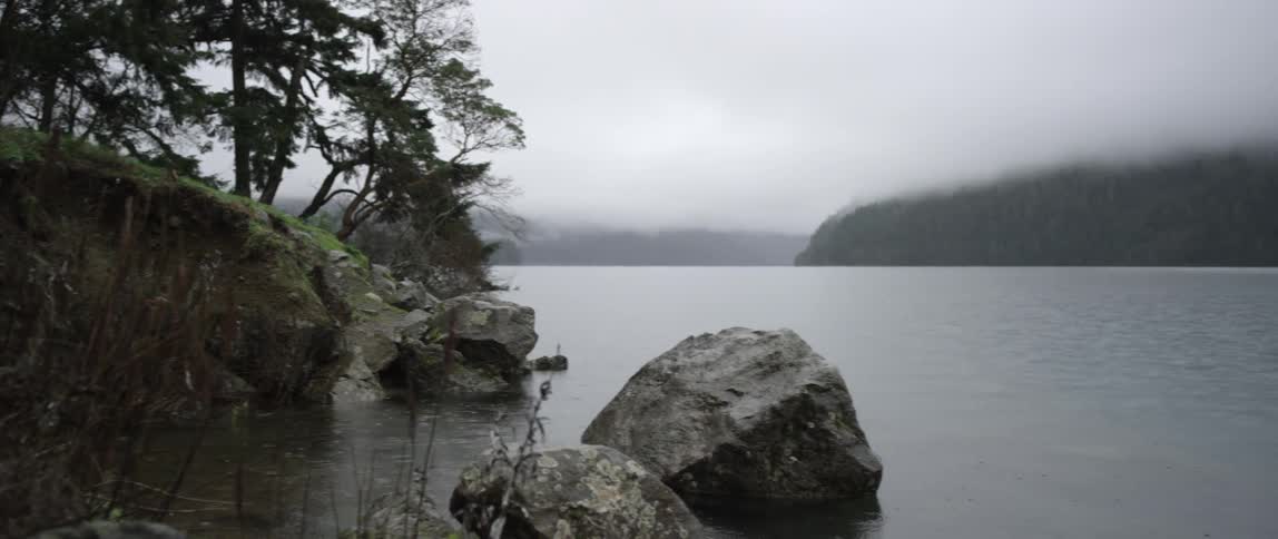View of a large lake with rocks at the shore and a large fog covered hill in the distance