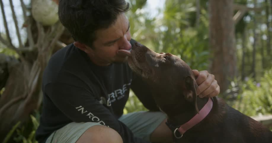 A man kneeling outdoors pets a dog as it licks his face