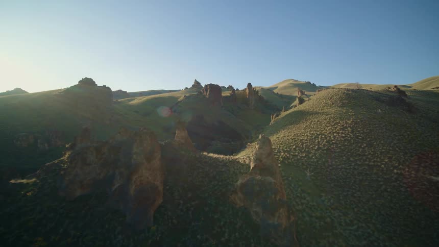Primeval spires and rock formations stand on rolling hills as the sun shines brightly on the horizon under a clear blue sky