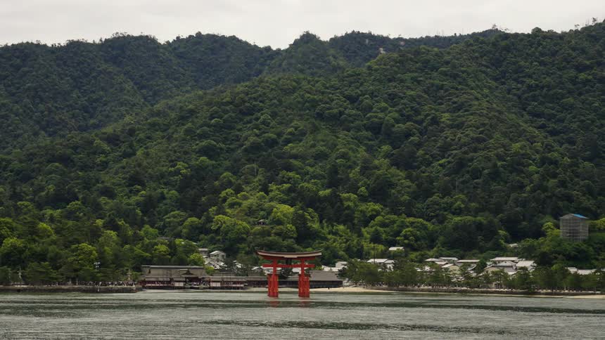 Water flows past torii arch standing in water a short distance from a Japanese town at base of a forest covered mountain