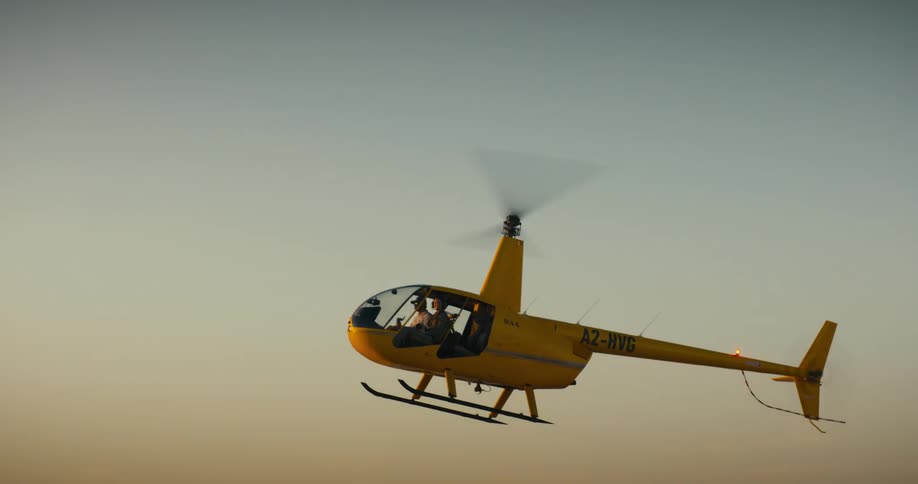 A yellow helicopter flies across a clear sky as it comes in for a landing