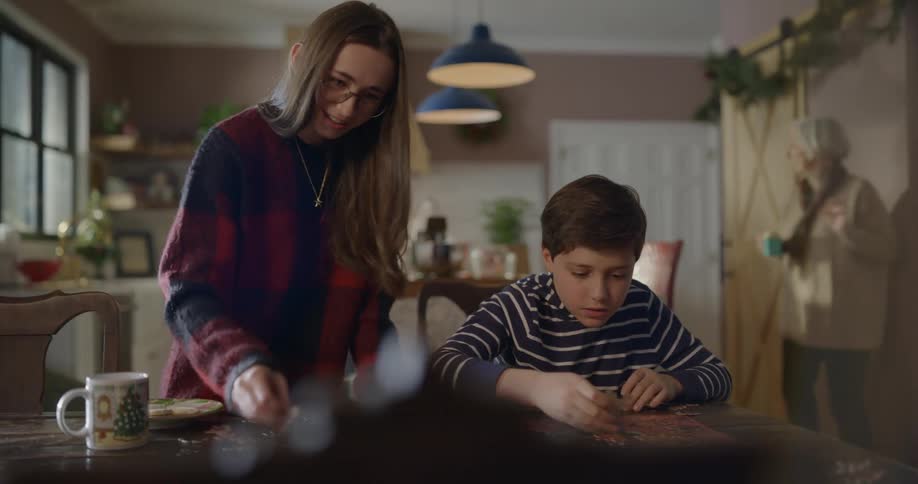 A teenage girl assembles a puzzle with a younger boy as two older women cook in the kitchen behind them