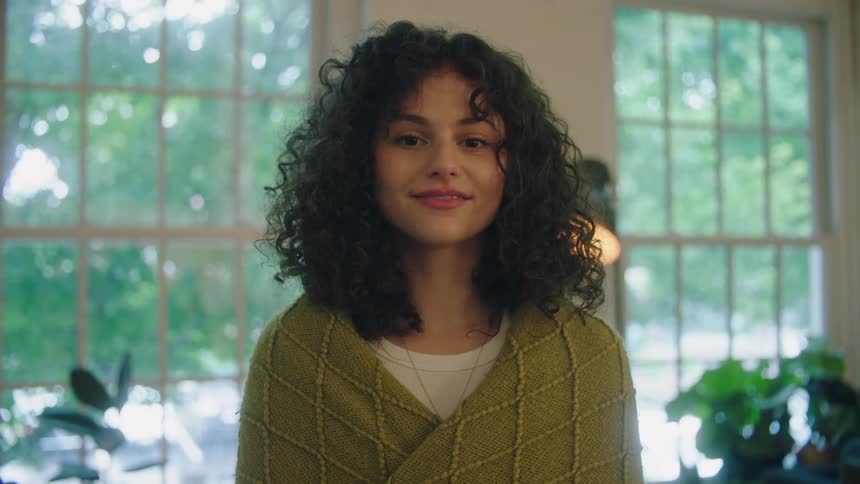 A young woman standing indoors with a blanket around her shoulders smiles into the camera