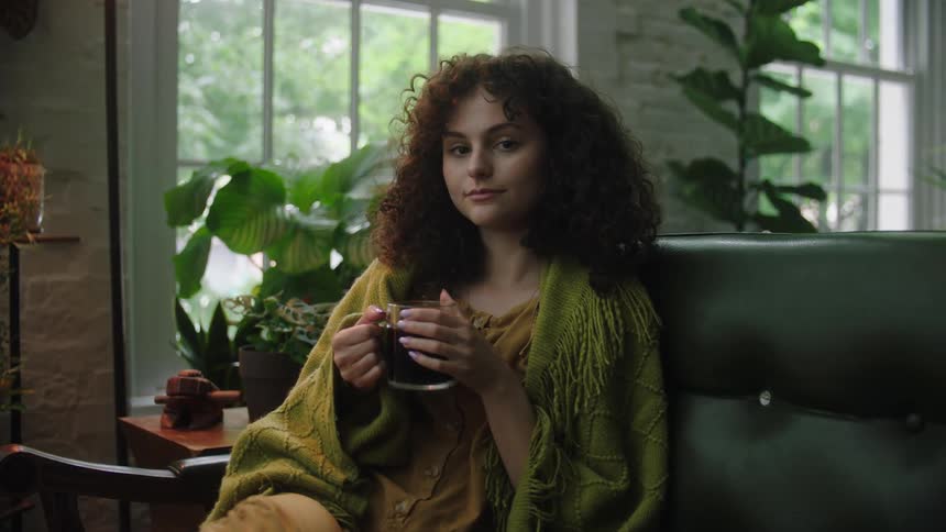 A young woman sits on a couch while holding a beverage in a cup