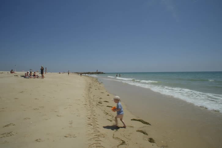 Child holding a toy bucket playing on the beach and running away from the gentle waves of the sea on a sunny day
