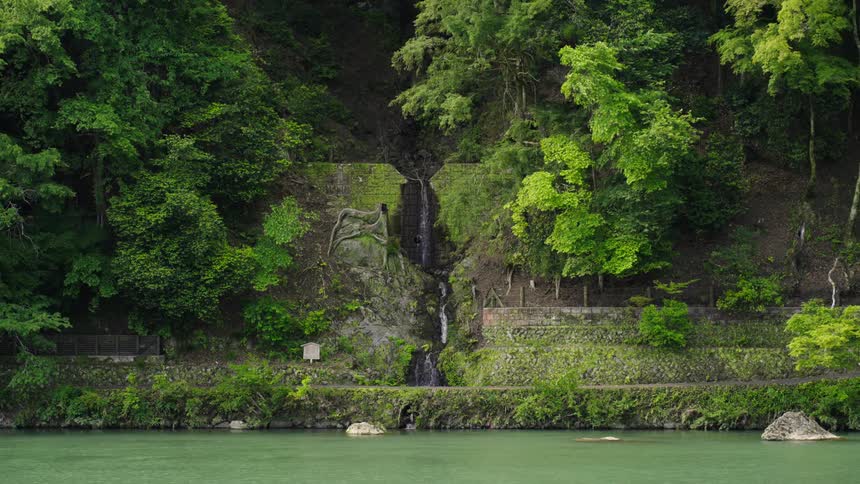 Narrow waterfall flows down steep mountainside past terraced manmade retaining walls toward green water