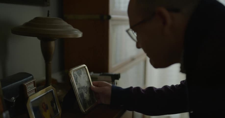A man looks at framed family pictures on a dresser and smiles