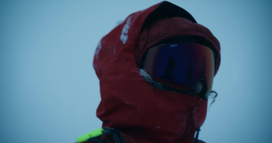 A woman wearing a red winter coat and goggles standing amidst a stormy weather condition