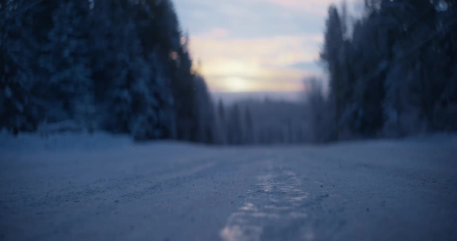 Snow and ice covered road leads through conifer forest to sun on red horizon