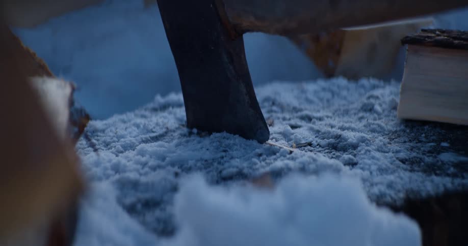 An axe is used to cut a piece of wood that was placed on a log of wood partly covered in snow in two more piece