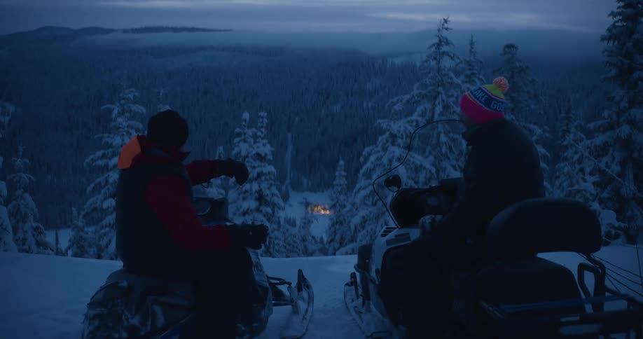 Friends sit on snowmobiles overlooking warmly lit building in clearing of snow covered conifer forest