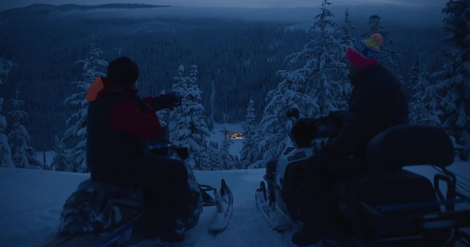 Male friends share drinks while sitting on snowmobiles overlooking snowy conifer forest surrounding a brightly lit house