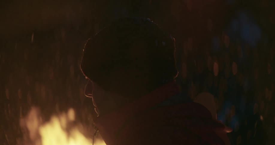 A man dressed for cold weather stands outdoors near a bondfire