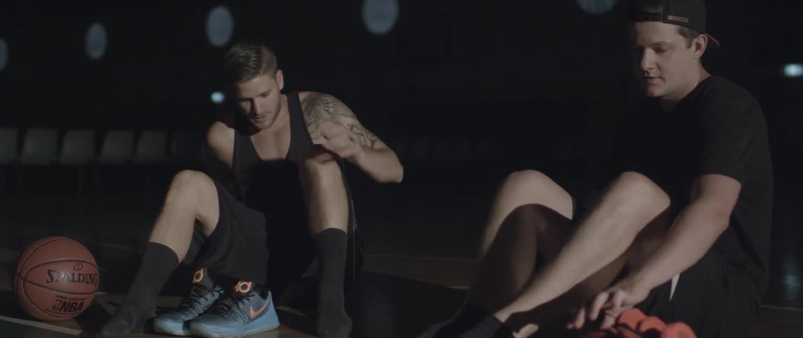 Two men carrying athletic shoes sit down on a basketball court to put their shoes on