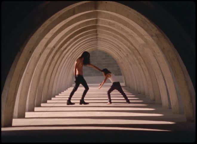 Tall shirtless man stops a child by the forehead inside an arched corridor