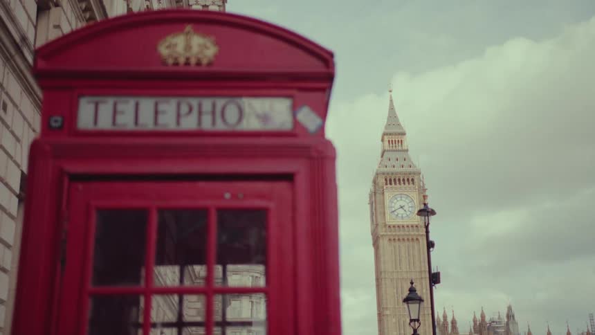 A red old fashioned telephone booth near a large clock tower outdoors