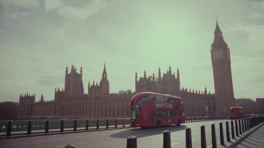 A group of people looking at a clock tower as red buses pass through the street