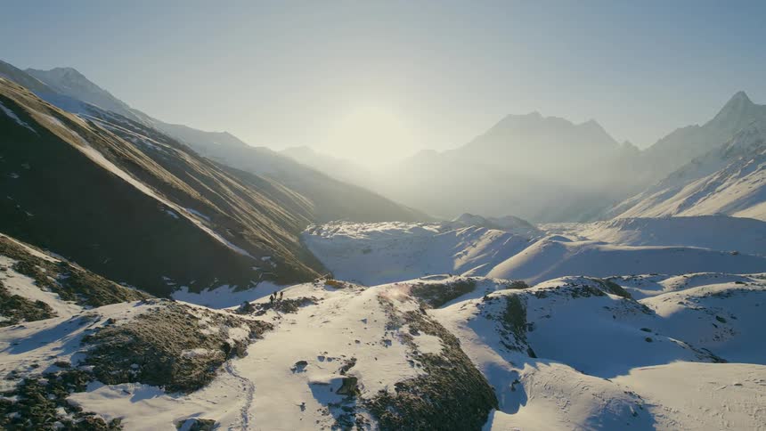People hike along trail over snow covered hills as sun shines from over mountains