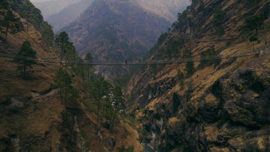 An aerial of a bridge connecting two mountains with a person walking over it and a river flowing underneath