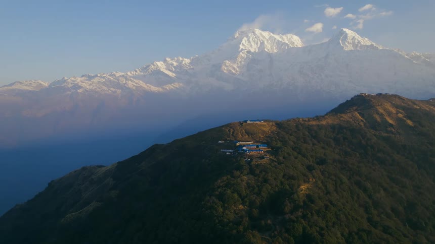 An aerial of a rural house on a forest covered mountainside with snow covered mountain ranges in the distance on a bright sunny day
