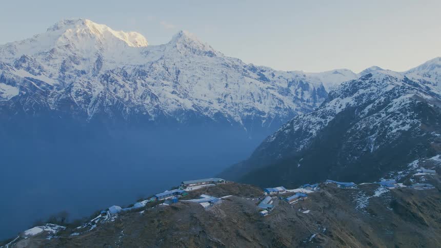 Tall snow covered mountains tower over town on greener slightly smaller mountain on foggy day