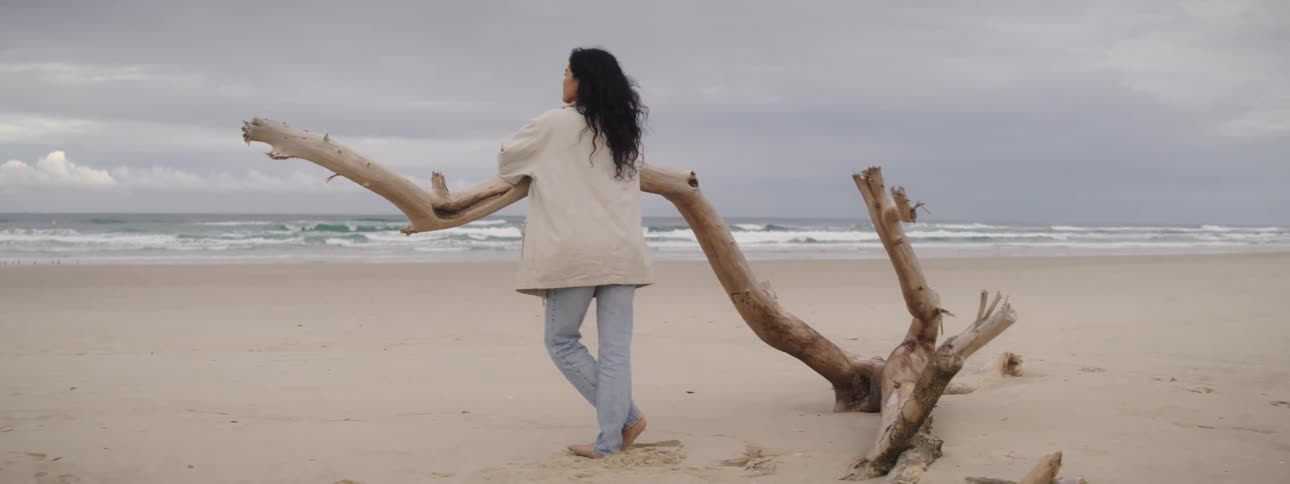 A woman leans on a large tree branch looking around at a beach on a cloudy day