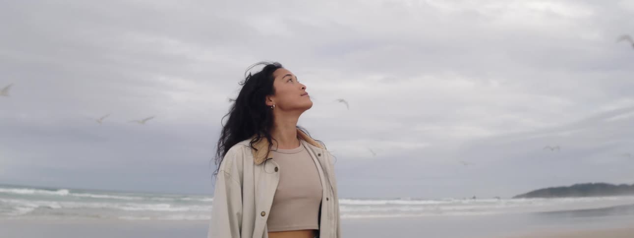 Woman standing on beach looks around as birds fly through overcast sky overhead