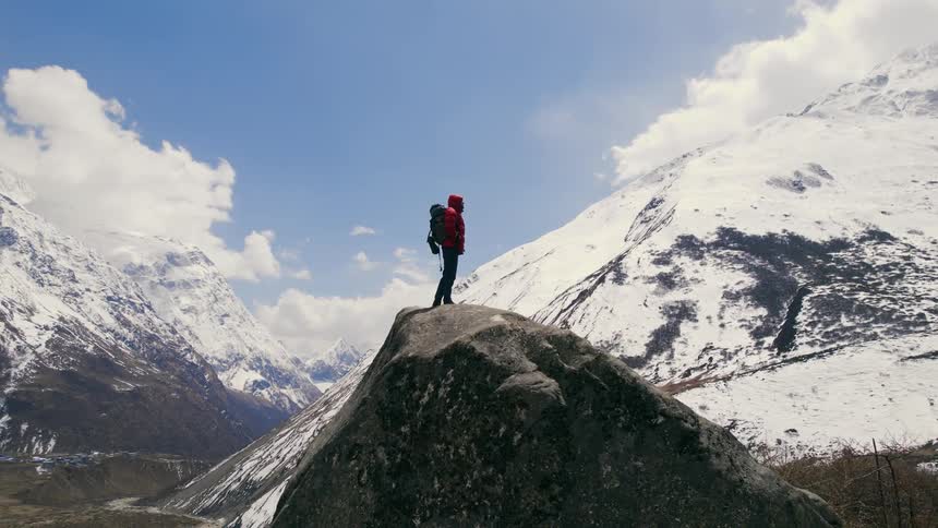 A man wearing a backpack is standing on a mountain peak looking at the surrounding mountains