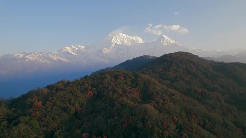 The sun shines upon forested mountains with trees in fall colors with snow covered mountains in the distance